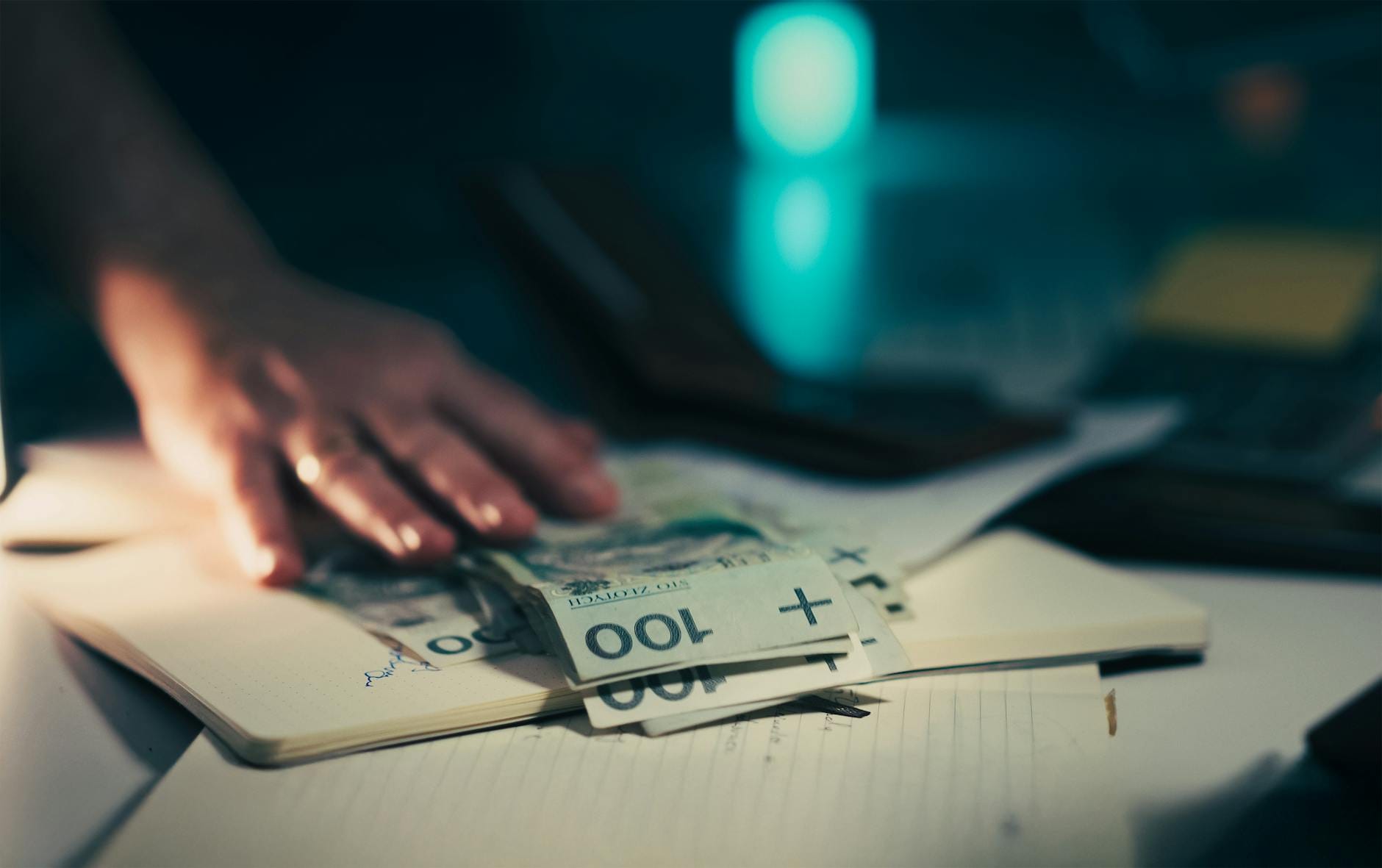 Dłoń sięgająca po banknoty na biurku obok notesu i kalkulatora
