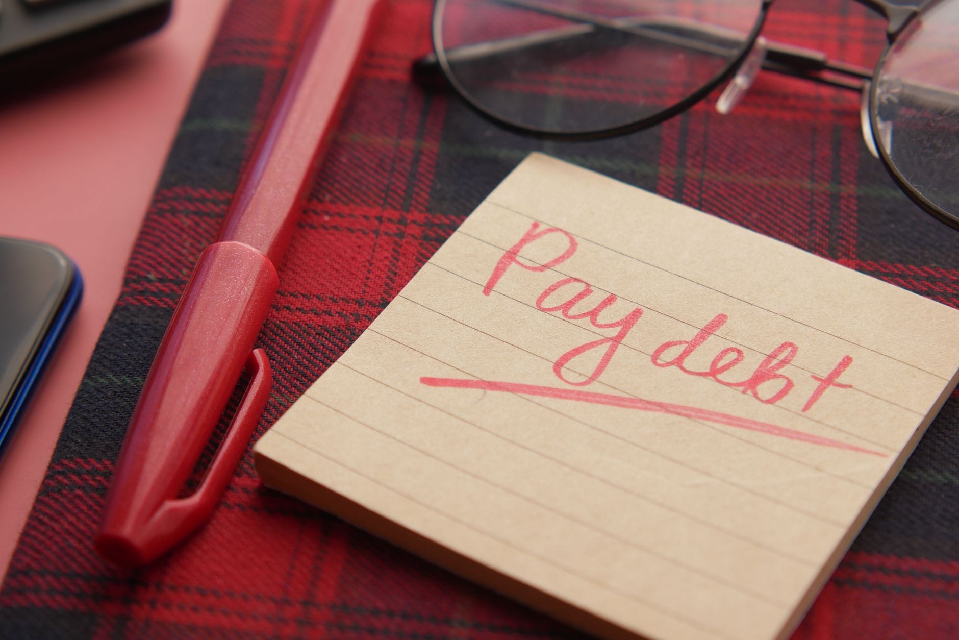 Kartka z napisem Pay debt i czerwony długopis na kraciastej tkaninie
